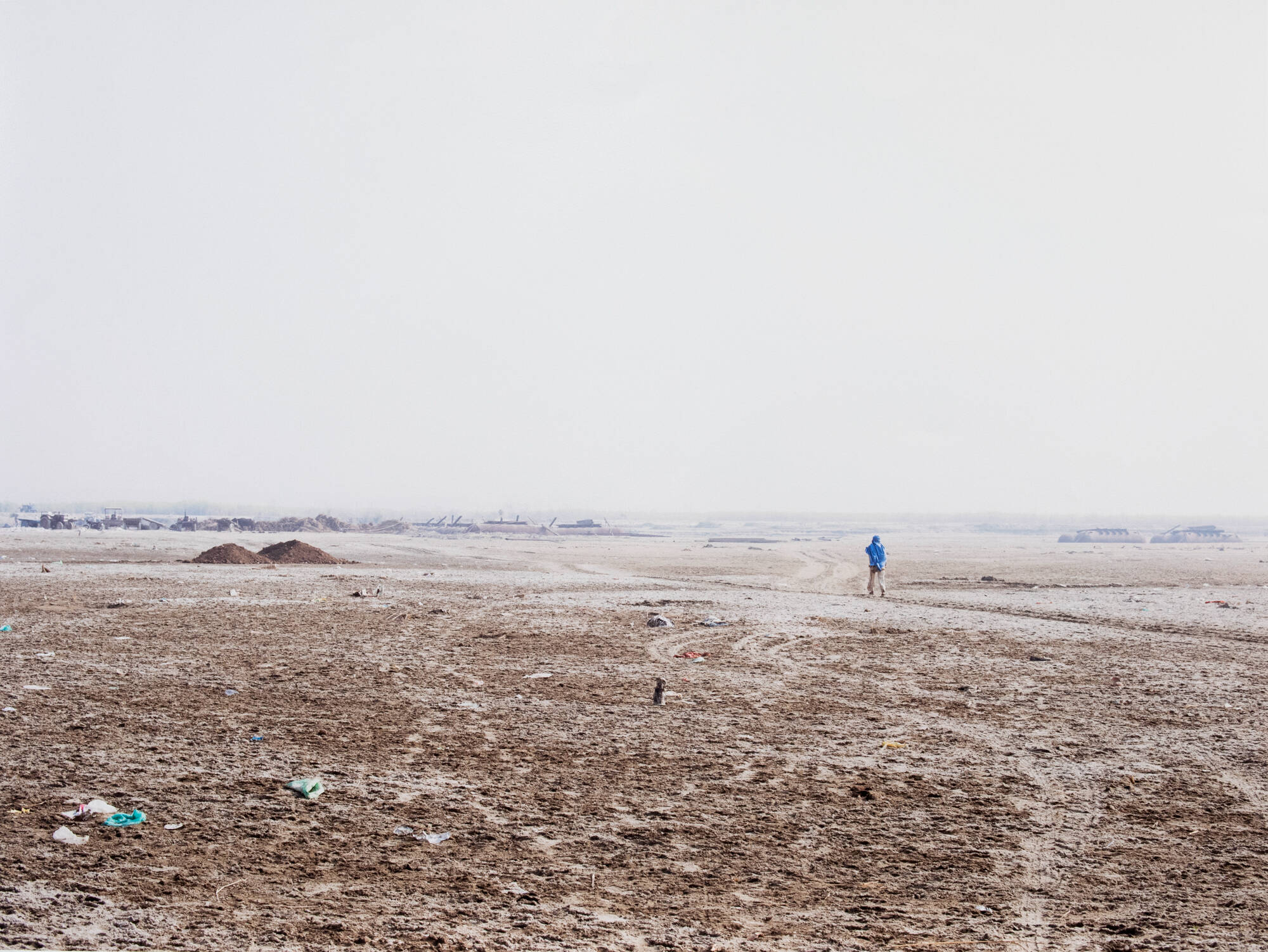 Color photograph of a desolate landscape with one person in the distance. Grey sky takes up half of the image, bottom of the image is mostly sandy with some water.